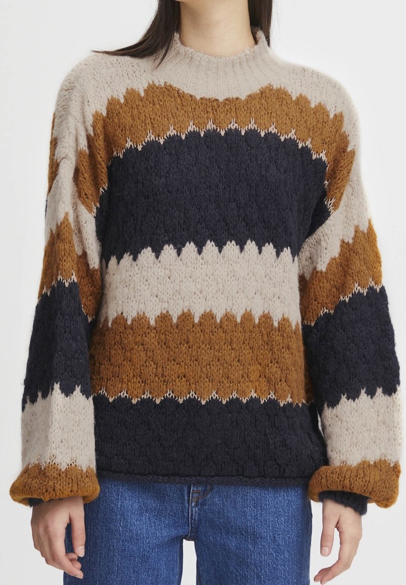 Pull en tricot épais avec de larges rayures horizontales en zigzag beige, marron et noir, porté avec un jean bleu, montrant le torse moyen et les bras.