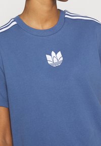 Blå bomullströja med vitt broderat logo på bröstet, rundad nacke och tre vita ränder på axlarna.