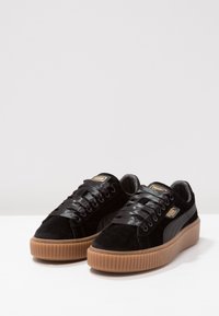 Svarta mockasneakers med platta svarta satinsnören, en strukturerad gumminsole och gyllene logotypaccenter på tungan och sidan. Plattformdesign.