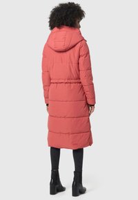 Lange pufferjas in koraal met een capuchon, elastische taille en gequilte ontwerp. Draagt over zwarte skinny jeans en enkellaarsjes.