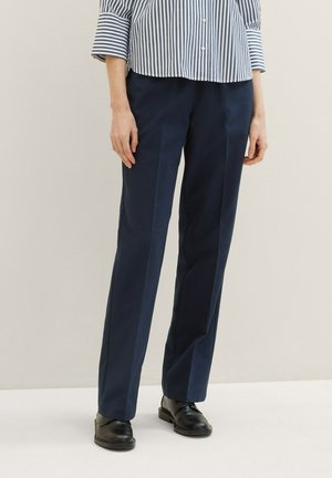 Pantalons taillés bleu marine avec une coupe droite et des plis avant visibles, associés à des chaussures en cuir noir. Le tissu semble lisse et structuré.
