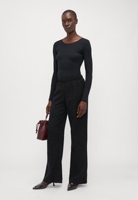 Black ribbed long-sleeve top paired with loose black textured trousers. Model holds a small burgundy bucket bag, wearing pointed black shoes.