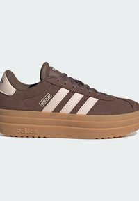 Smeđe nubuk tenisice s tri bijele pruge, zaobljenim prstom, debelim gumiranim potplatom i brendiranim detaljima sa strane i na jeziku.