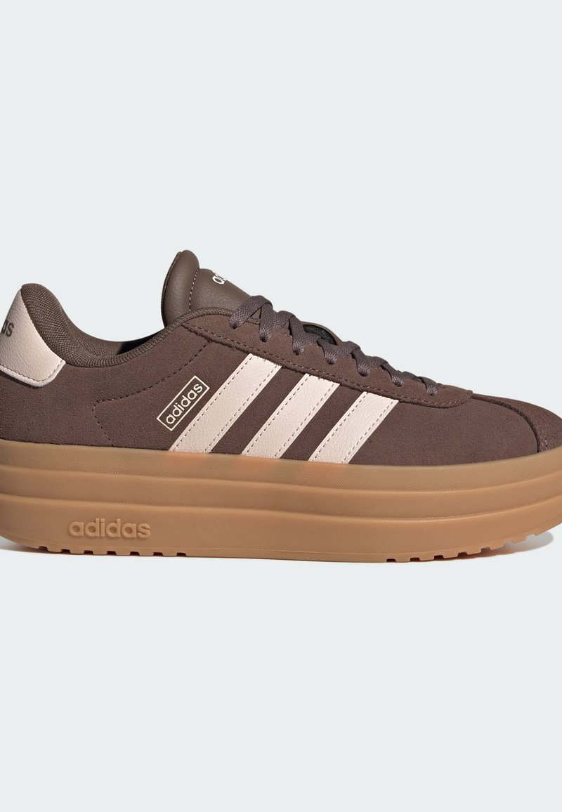 Bruine suède sneakers met drie witte strepen, ronde neus, dikke gumzool en merkaccenten aan de zijkant en op de tong.