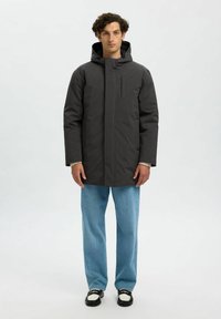 Giacca impermeabile nera con cappuccio, zip frontale e tasca sul petto. Indossata su jeans azzurri chiari e scarpe nere, presenta un fit rilassato.