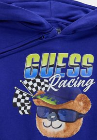 Koningblauwe hoodie met een afbeelding van een beer die een zonnebril en een hoed met vlammen draagt, naast racenvlaggen en de tekst "GUESS Racing".