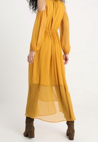 Długa żółta sukienka z lekkiego materiału, z przezroczystą dolną częścią, otwartym dekoltem na ramionach, elastycznym pasem i zwiewnymi rękawami. Czekoladowobrązowe, do kolan sięgające botki.