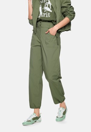 Person wearing olive green cargo pants with elastic cuffs, matching jacket, graphic tee, and light green and white sneakers, standing with hand in pocket.