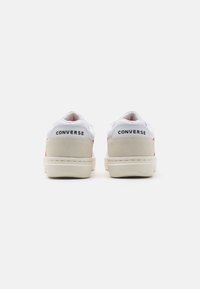Adidași din piele albă cu accente roșii, toc din daim, talpă din cauciuc și logo-ul negru "CONVERSE" pe spate, având un design minimalist.