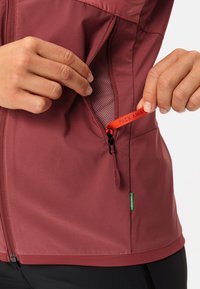 Giacca bordeaux con una tasca con zip, dotata di una fodera in rete, un cinturino rosso a contrasto con la scritta "PACK AWAY" e un'etichetta verde all'orlo.