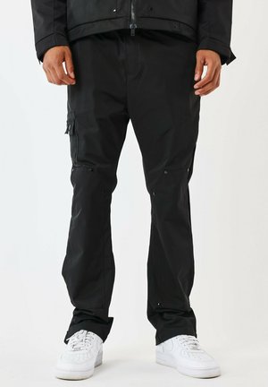 Pantalons imperméables noirs avec une texture lisse, dotés de poches cargo, de fermetures à pression et d'un design à jambes droites. Portés avec des baskets blanches.