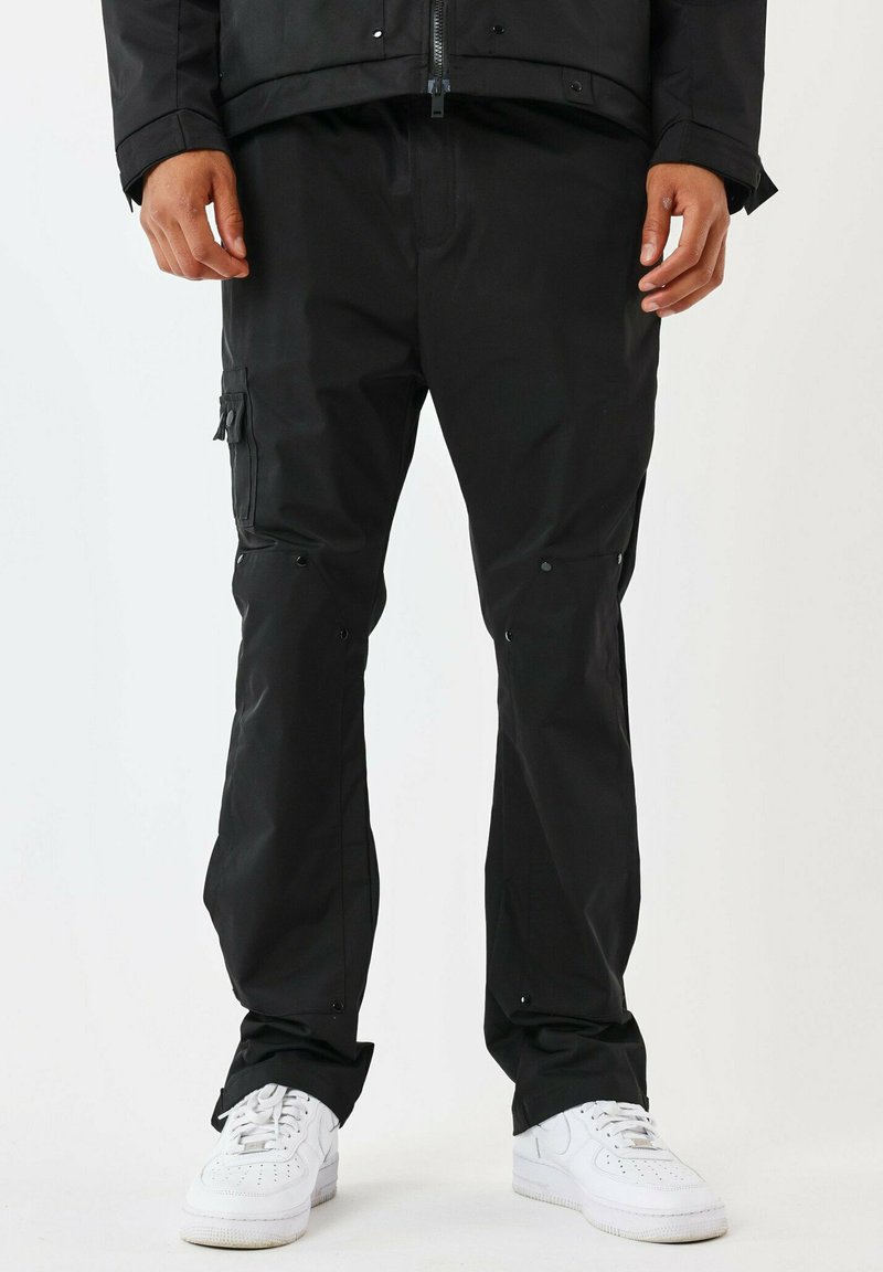 Pantaloni neri impermeabili con una texture liscia, dotati di tasche cargo, chiusure a scatto e design a gamba dritta. Indossati con sneakers bianche.
