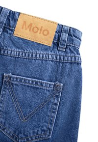 Denimo audinys mėlyno atspalvio turi smėlio spalvos odinį etiketę su įspaudu "Molo". Gale esančioje kišenėje matyti išskirtinis V formos siuvinėjimo raštas.