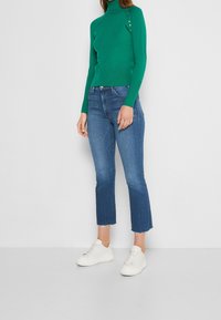 Grön polokrage tröja med knapppdetails, tillsammans med avkortade blåjeans och vita sneakers. Outfitten har en åtsittande silhuett.