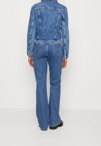 Denim jacket and flared jeans in medium blue, featuring a fitted silhouette, prominent seams, and two back pockets with button closures.