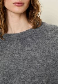 Siv volnen pulover z rebrastim ovratnikom in mehko, teksturirano površino. Funkcija bližnjega posnetka poudarja njegovo toplo, udobno blago.