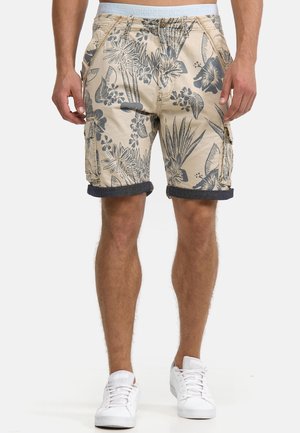 Mand iført beige cargoshorts med mørkeblå tropisk blomsterprint og opkrogede kanter, kombineret med hvide sneakers og synligt gråt linning.