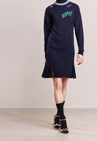 Abito lavorato a maglia navy con colletto a coste e pieghe laterali, caratterizzato da dettagli ricamati. Indossato con calze nere e sandali con tacco con bordi decorativi.
