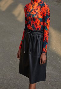 Modell som bär en livfull röd, blå och orange blommig långärmad topp instoppad i en svart läderkjol med dragsko i midjan och stora fickor.