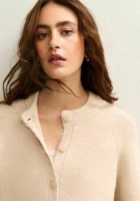 Cárdigan beige con un tejido texturizado, cuello redondo y cinco botones en la parte delantera. Presenta mangas raglán y un acabado suave.