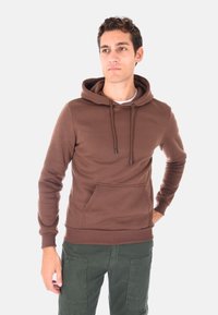 Uomo che indossa una felpa con cappuccio marrone e pantaloni cargo verdi, in piedi con una mano sui fianchi, che guarda leggermente di lato.