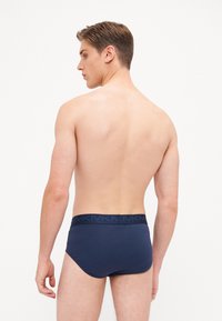 Caleçons en coton bleu marine avec une ceinture flexible mettant en valeur une bande plus foncée et un détail de logo, présentés sur un modèle masculin de dos.