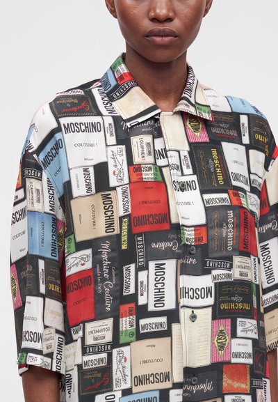 Gros plan d'une personne portant une chemise multicolore recouverte de différentes impressions d'étiquettes de la marque Moschino dans diverses couleurs et polices.