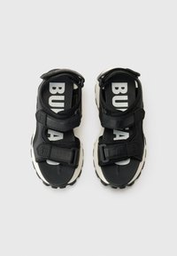 Chinelos pretos com tiras ajustáveis, tecido texturizado, letras brancas nas palmilhas e uma sola grossa branca com um padrão negro rugoso.