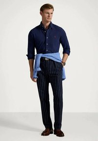 Camisa azul-escura de botões com colarinho, combinada com calças azul-escuro riscada. Sweater azul claro amarrado à cintura, sapatos castanhos.