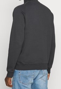 Persona vestida con un sudadera negra de manga larga y jeans de denim azul claro, mostrada de espaldas con una mano relajada a un lado.