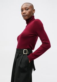 Burgundi turtleneck pulóver, melyet magasan szabott fekete nadrággal párosítottak, arany csattal. Simaságot sugalló, kötött textúra, alakhoz illeszkedő ujjak.