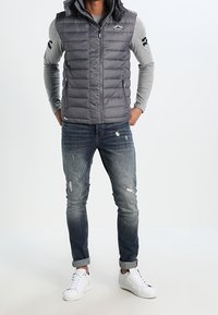 Grå pufferväst med huva, zip-förslutning framtill och sidfickor, bärs över en grå långärmad skjorta och slitna blå jeans med uppvikta ärmslut.