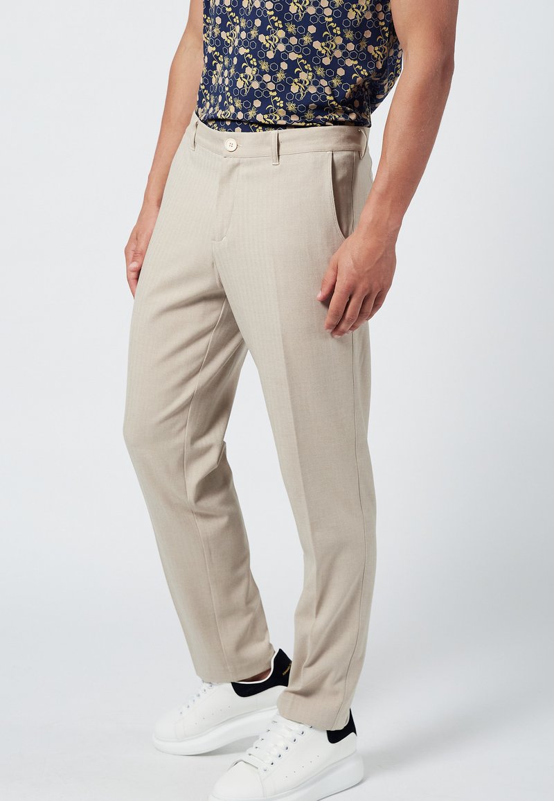 Pantalons sur mesure beiges avec une finition texturée, dotés d'un devant plat, de poches latérales et d'une fermeture à un bouton. Associés à des baskets blanches.