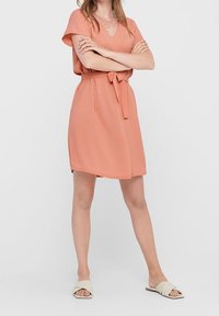 Persiko färgad klänning med korta ärmar, V-ringning och midjeband. Slät tyg, knälång med sidofickor, bärs med lätta sandaler.