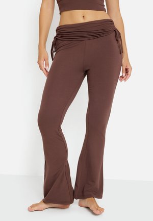 Vrouw draagt bruine, hoog getailleerde wijde broek met gerimpelde koordsluiting in de taille, staat blootsvoets tegen een effen witte achtergrond.