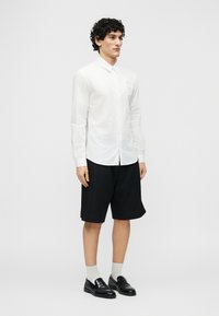 Νεαρός άνδρας όρθιος, φοράει λευκό πουκάμισο με μακριά μανίκια, μαύρα σορτς μέχρι το γόνατο, λευκές κάλτσες και μαύρα μοκασίνια, μπροστά σε απλό φόντο.