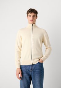 Pull zippé crème avec col haut, ourlet côtelé et poignets assortis, arborant un logo discret sur la poitrine gauche. Porté avec un jean bleu.
