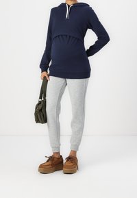 Marineblauwe hoodie met een getailleerde voorkant en trekkoord, gecombineerd met grijze joggingbroek. Model draagt bruine plateau schoenen en houdt een groene tas vast.