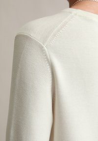 Helle beige Strickjacke mit strukturiertem Gewebe, Rundhalsausschnitt und Raglanärmeln, die dezente Nähte an den Schultern zeigt.