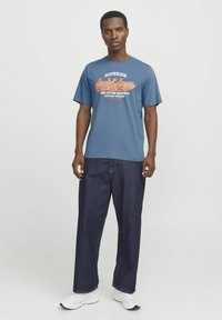 Blauwe katoenen T-shirt met de tekst "SUPERIOR JACK & JONES" in oranje en wit, gecombineerd met donkerblauwe wijdegemeten jeans en witte sneakers.