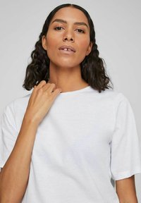Tricou alb cu mânecă scurtă și guler rotund, purtat de o persoană cu păr creț. Materialul pare neted și ușor.