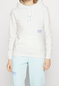 Sudadera blanca de algodón con bolsillo canguro, detalles en verde menta y logo. Combinada con pantalones de chándal en verde menta que tienen logos en cada uno.