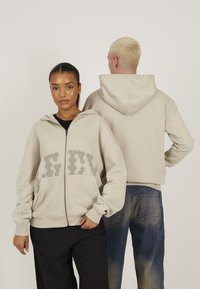 Beige mikina s kapucňou na zips a text�úrovaným logom vpredu, s kapucňou, dlhými rukávmi a voľným strihom. Zadný pohľad zobrazuje jednoduchý dizajn.