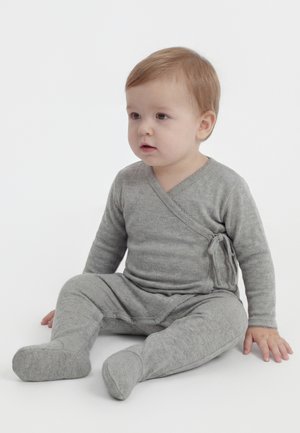 Νήπιο με ανοιχτό καστανά μαλλιά καθισμένο, φοράει γκρι υφασμάτινη πλεκτή μπλούζα τύπου τύλιγμα και αντιστοιχων ποδιών παντελόνια, σε λευκό φόντο.