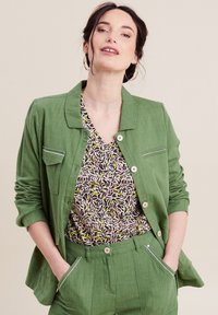 Giacca di lino verde con colletto e tasca frontale, abbinata a una blusa con motivo caratterizzato da spirali di nero, rosa e giallo.