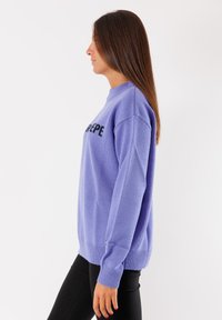Maglione viola chiaro con scollo rotondo, spalle scese e scritte nere. Presenta polsini e orlo a coste, realizzato in tessuto a maglia testurizzato.