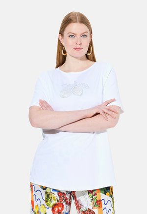 Bílý tričko s krátkým rukávem, které na hrudi zdobí design ovoce posetý rhinestonem, v kombinaci s barevnými kalhotami s květinovým vzorem.