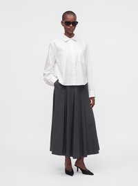 Femme portant des lunettes de soleil noires, une chemise blanche boutonnée, une longue jupe plissée gris foncé et des chaussures noires à talons, debout devant un fond uni.