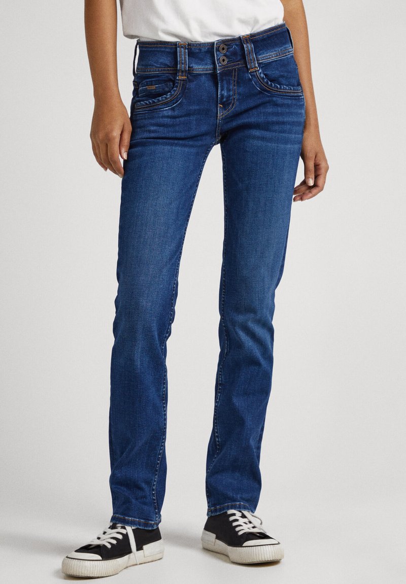Jeans de mezclilla azul con una cintura ajustada, cierre de dos botones, bolsillos frontales con costuras curvadas y piernas rectas, combinados con zapatillas negras.