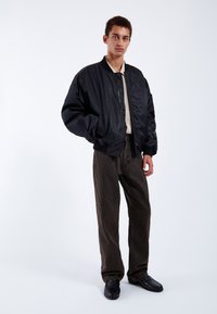 Black bomber jacket with a shiny finish, ribbed cuffs, beige shirt underneath, brown pants, black shoes, and a minimalist backdrop.
